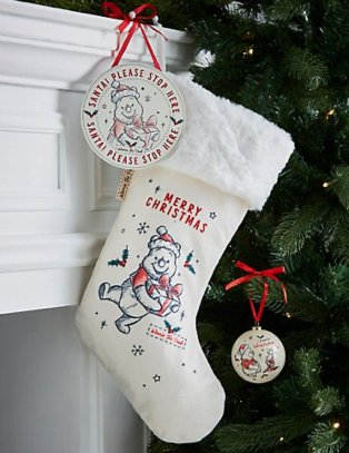 Cream Winnie the Pooh stocking with white faux fur trim hangs from mantelpiece with Christmas tree in the background.