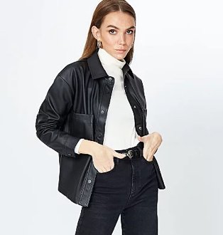 Brunette woman poses wearing white roll neck top, black leather-effect shacket and black belted jeans.