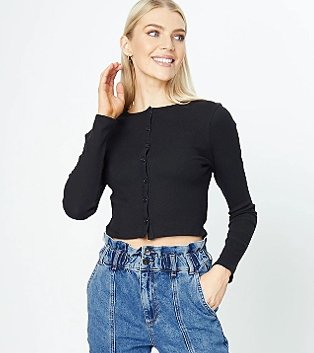 Blonde woman poses smiling raising hand to face wearing black knit cropped length button down cardigan and blue paper bag jeans.