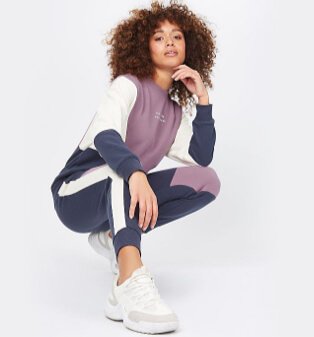 Woman poses crouched down wearing grey, purple and white hoodie and trousers.