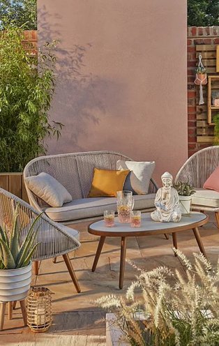 Patio with nerja 4 piece garden sofa set and matching table topped with a glass jug, two glasses and a buddha ornament surrounded by artificial plants.