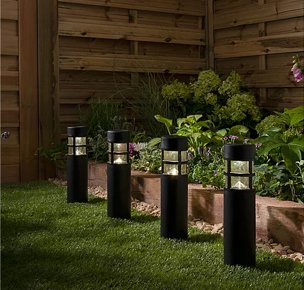 Fenced garden with a line of 4 black outdoor white LED lighthouse pillars placed on grass with an array of plants and greenery in the background.