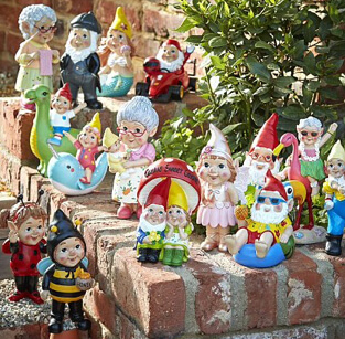 A large collection of garden gnomes on a garden wall, including one dressed in a bumblebee costume, one riding a dinosaur and more