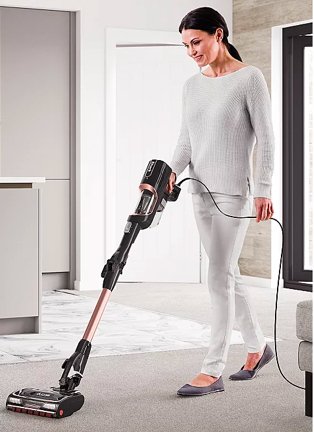 Woman vacuums grey carpet wearing grey lounge top and bottoms and dark grey ballet shoes.