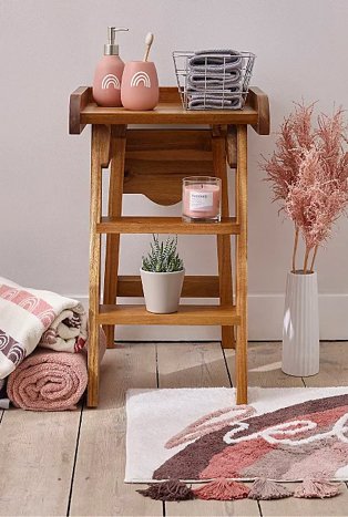 Wooden table topped with pink rainbow dispenser and tumbler set, grey storage basket, pink candle and artificial plant surrounded by pink and cream rolled towels, pink artificial plant in white vase and cream, brown and pink hello slogan bath mat.