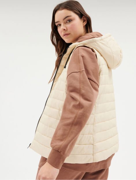 A woman posing in a cream padded gilet over a brown hoodie and joggers set.