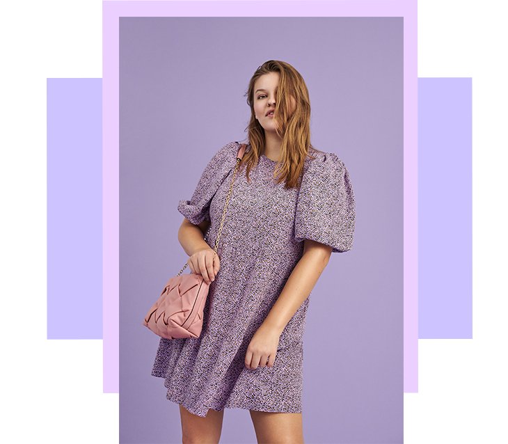 Woman poses smiling wearing purple floral puff sleeve dress with pink quilted shoulder bag.
