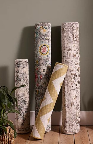4 assorted abstract printed rugs stood upright on wooden floor against a grey wall.