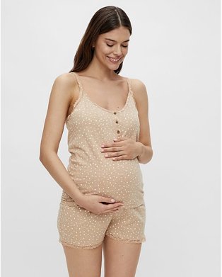 Woman smiles looking down cradling bump wearing beige spotty maternity vest top and shorts pyjama set.