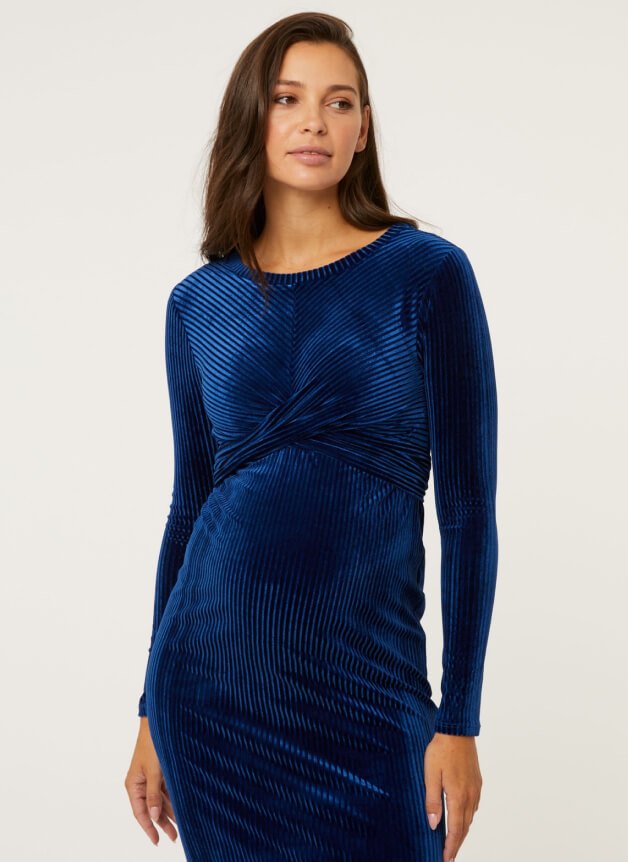 Woman poses wearing maternity blue velvet ribbed wrap around dress.
