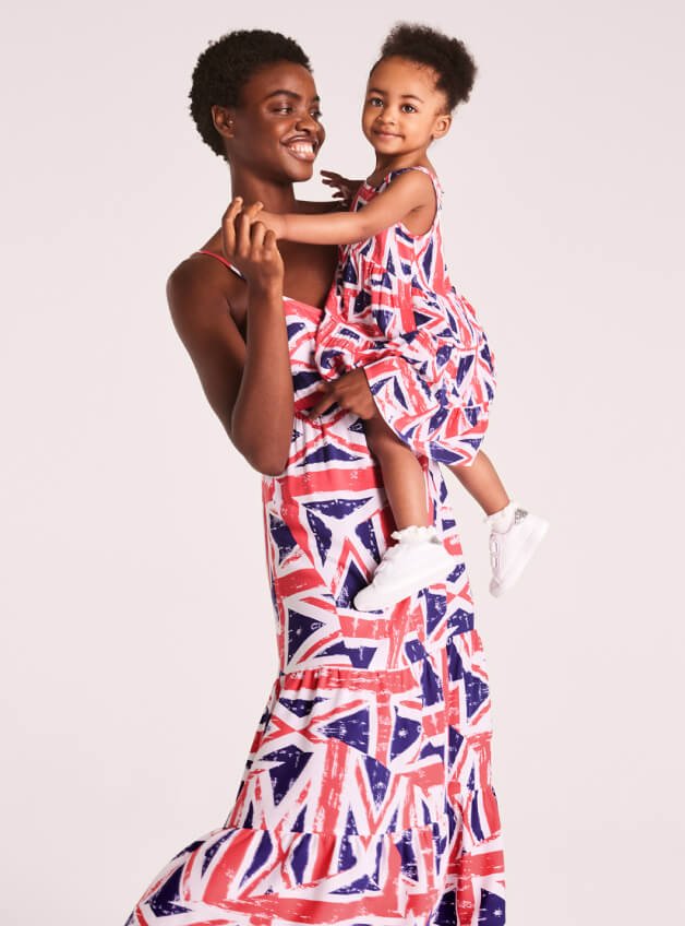 A woman smiles holding a child both wearing matching Coronation Union Jack flag midi sundresses.