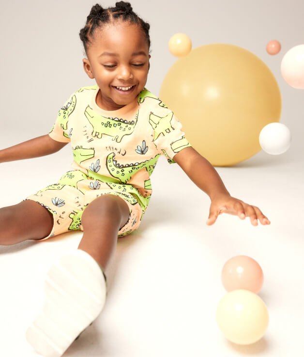 Boy smiles while playing with balls wearing orange and green dinosaur printed shorts and t-shirt outfit.