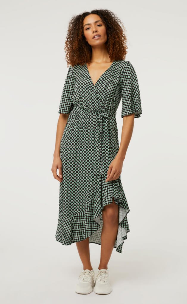 Woman poses wearing green and black polka dot midi dress and white pumps.