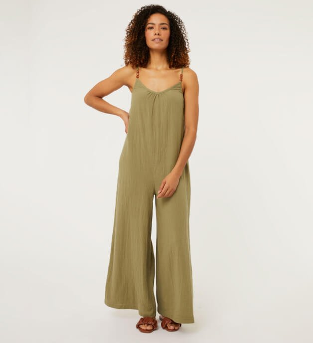 Woman poses with hand on hip wearing a khaki bead strap lightweight jumpsuit.