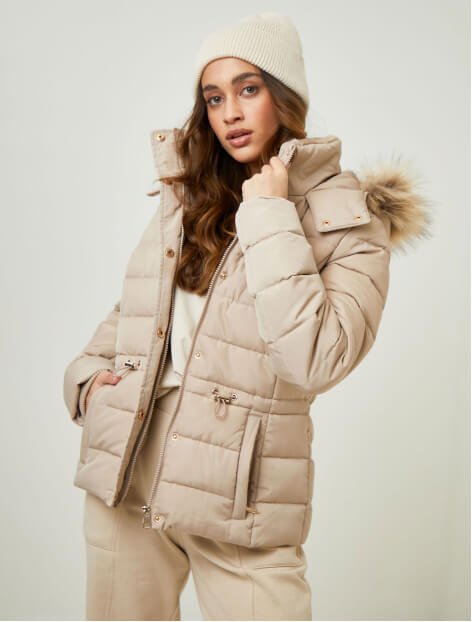 A woman posing in a beige faux fur trim padded coat, beige trousers and beige banie hat.