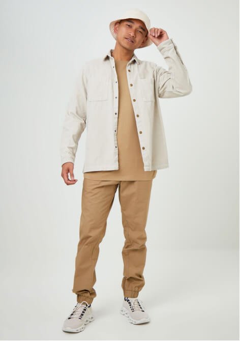 A man posing in a beige joggers and t-shirt set, cream jacket, cream hat and neutral trainers.
