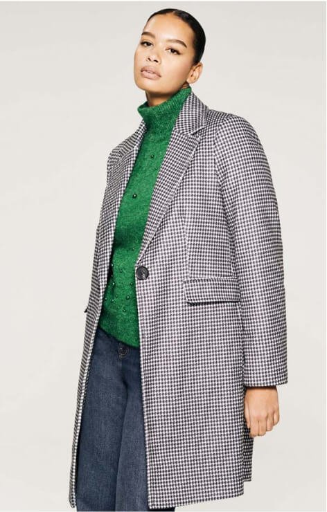 A woman wearing a mono houndstooth coat over a green knitted jumper and dark wash jeans.