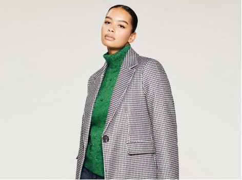 A woman wearing a houndstooth coat over a green roll neck jumper and jeans