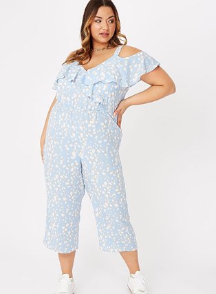 Woman poses with one hand on hip wearing blue abstract spot print cold shoulder jumpsuit and white trainers.