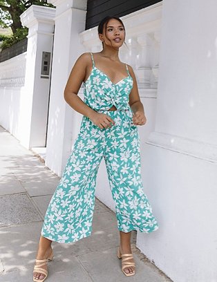 Woman poses with hands on hips in front of large white wall wearing green floral print twist front jumpsuit and nude heeled sandals.