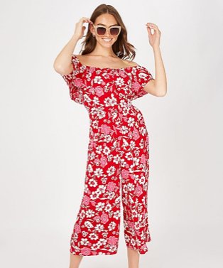 Woman poses smiling with hands raised to head wearing red floral cold shoulder jumpsuit and nude oversized sunglasses.