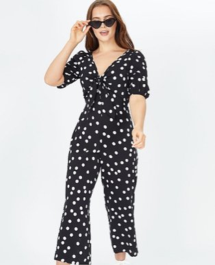 Woman poses holding side of sunglasses wearing black polka dot print jumpsuit.