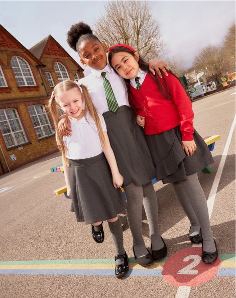 Three school children in a playground wearing a selection of school uniform items including grey skirts, white shirts, ties and cardigans.