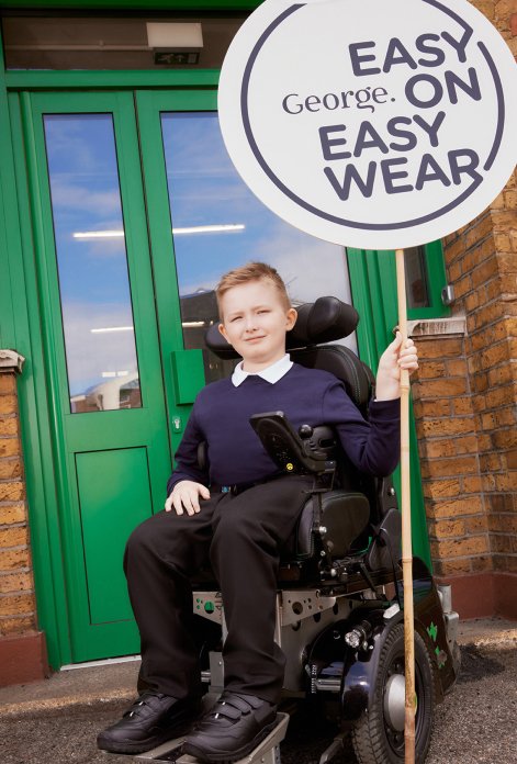 A child in a wheelchair wearing black trousers, black shoes, a white polo shirt and navy jumper holding a 'George Easy On Easy Wear' sign.