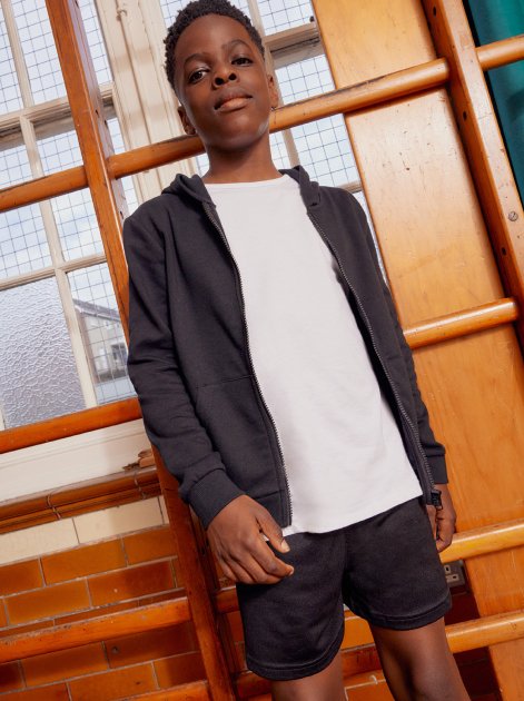 A boy wearing a white t-shirt, black shorts and jacket in a school gym.