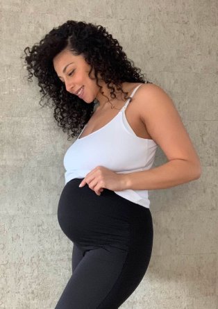 Pregnant woman poses with hands on bump wearing white cami top and black over the bump leggings.