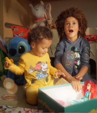 Two children wearing Disney jumper and joggers outfits look amazed opening box with Disney plush toys in the background.