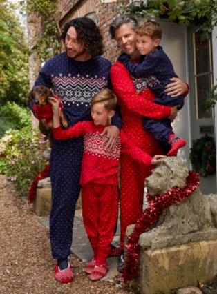 Two men, two children and a sausage dog huddle together wearing matching red and blue festive Fairisle outfits with greenery in the background.