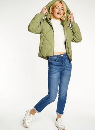 Woman poses lifting hood of jacket wearing cream knitted jumper, khaki hooded padded jacket, mid-wash blue jeans and white trainers.