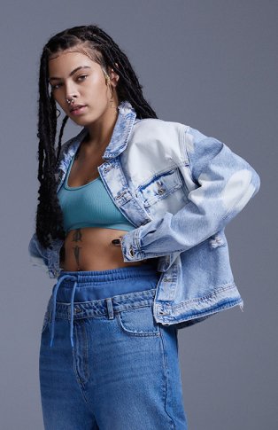 Woman poses side-on wearing mid-blue jogger jeans, turquoise crop top and acid wash denim jacket.