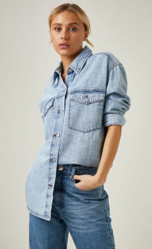 Woman poses with one hand in pocket wearing light denim button-down shirt tucked into mid-blue jeans.
