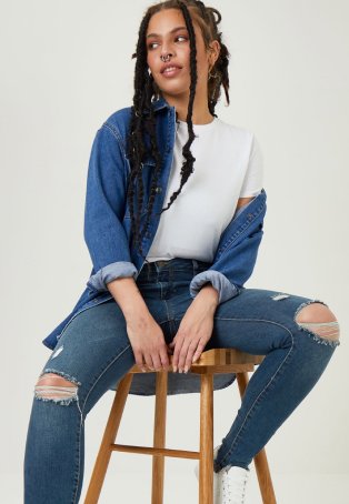 Woman sits on wooden stool wearing white tee, blue shacket and mid-blue distressed jeans.