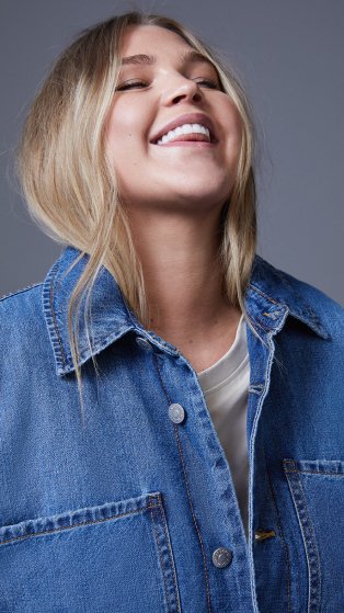 Woman tilts head back smiling wearing a blue mid-blue oversized denim jacket.