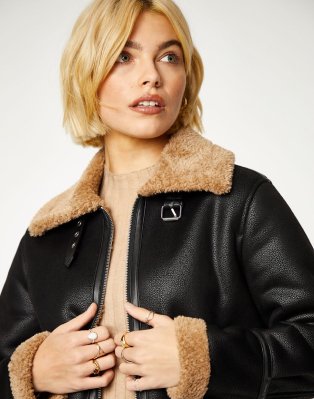 Woman poses wearing tan knitted jumper dress and black faux-leather jacket with faux-fur collar and cuffs.